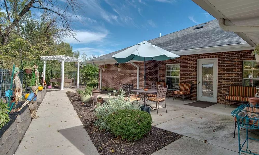 A cozy patio area with outdoor seating and an umbrella, adjacent to a brick building surrounded by well-maintained landscaping.