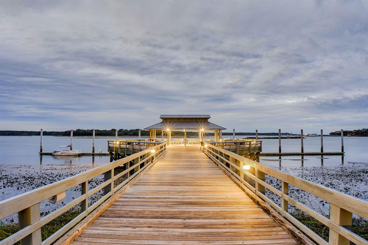 A wooden pier illuminated by warm lights stretches over a calm body of water under a cloudy evening sky.