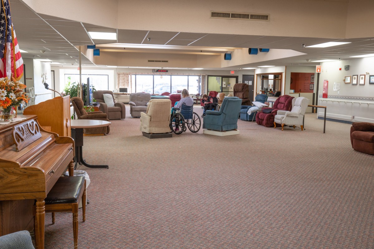 A spacious lounge area with various chairs and a wheelchair on a carpeted floor, illuminated by large windows.