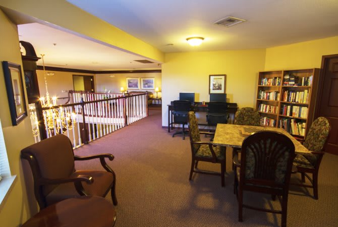 A cozy library interior featuring bookshelves, a reading area with tables and chairs, and computers under warm lighting.