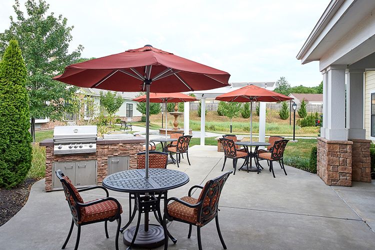 An outdoor patio area with tables, chairs, and red umbrellas in a garden setting.