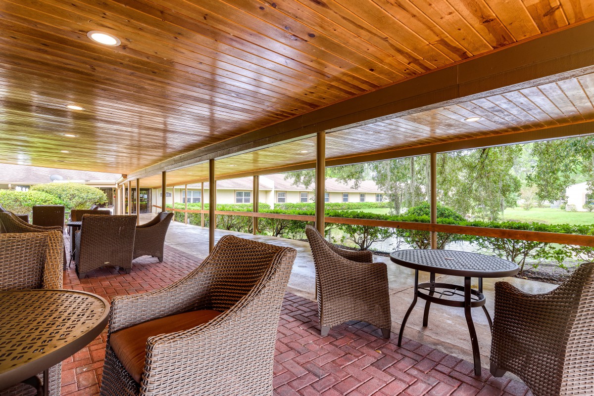 A cozy outdoor patio area with wicker furniture and tables, set on a brick floor with a wooden ceiling, overlooking a green landscape.