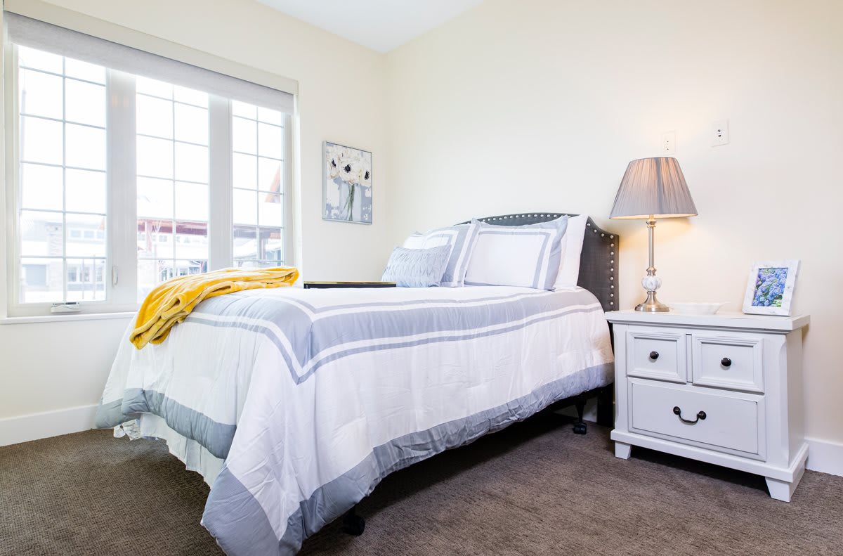 A cozy bedroom with a neatly made bed, featuring a bedside lamp and nightstand next to a large window.