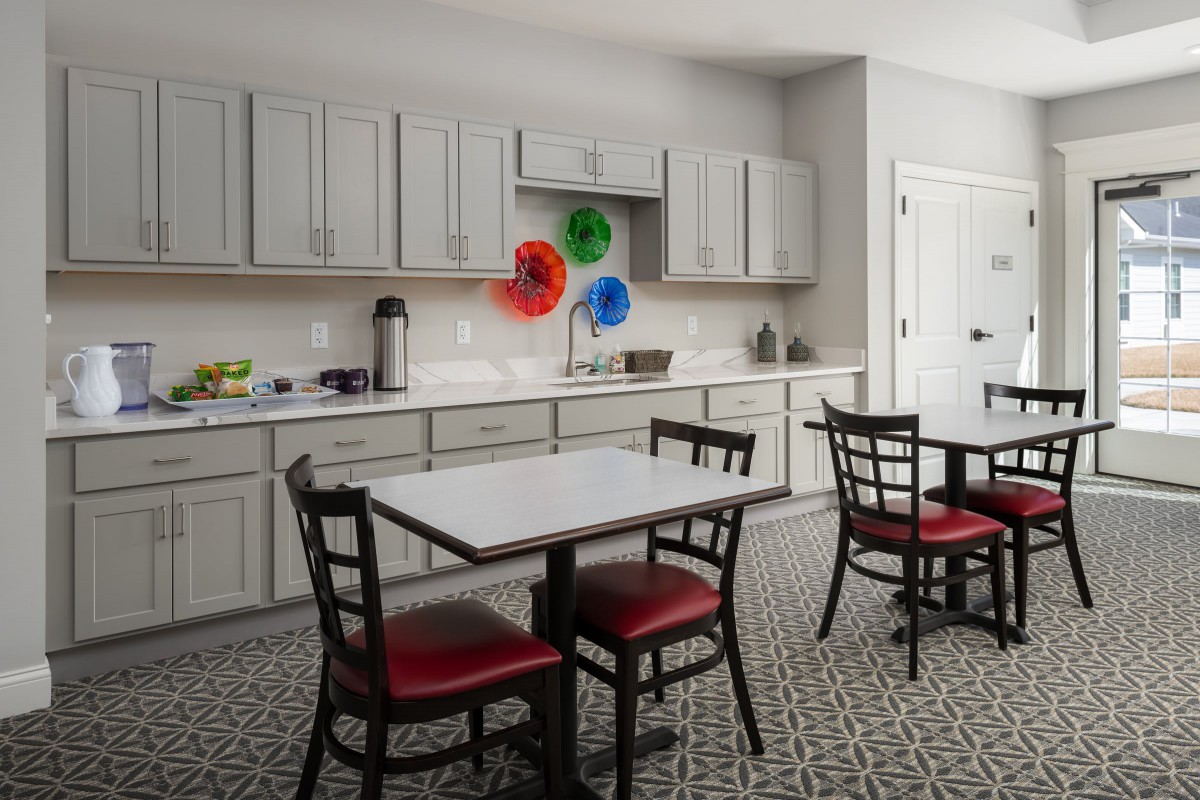 A modern kitchen with a dining area featuring red cushioned chairs and a small table, complemented by neutral cabinets and colorful wall decorations.