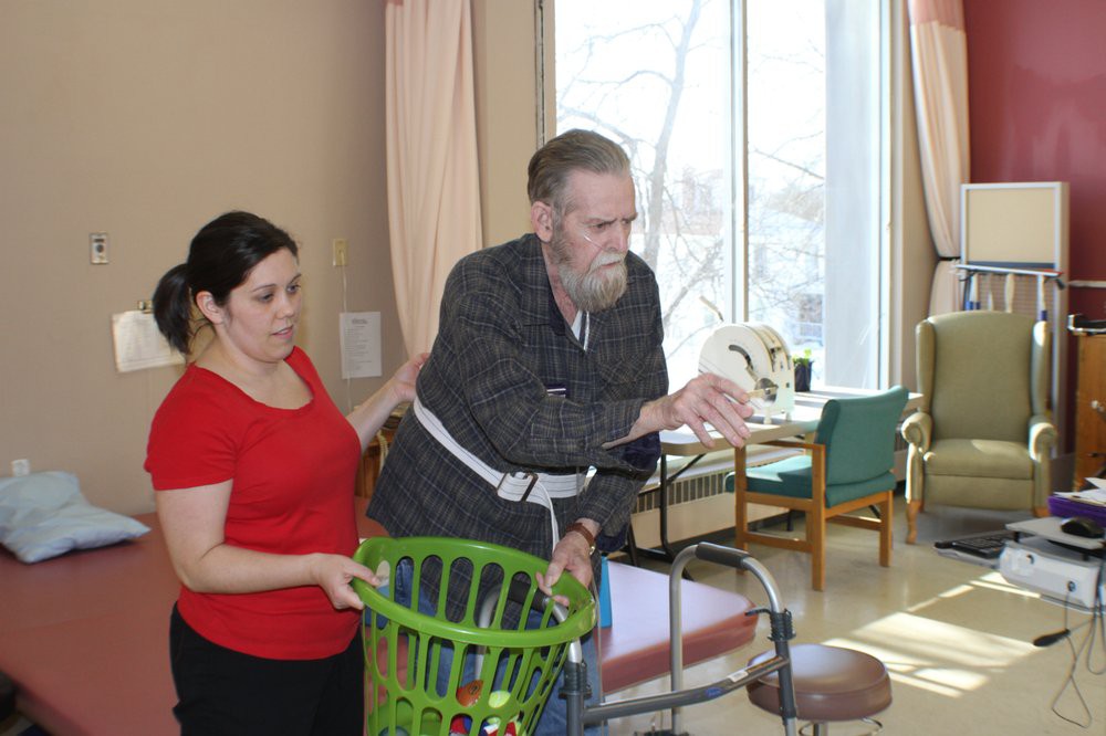 A caregiver assists an elderly man using a walker in a rehabilitation setting.