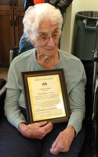 An elderly woman proudly holding a certificate in a warm indoor setting.