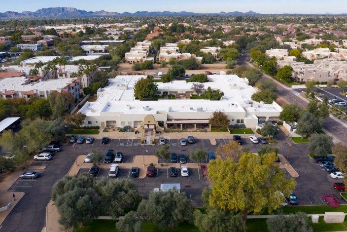 Aerial view of a building complex with white roofs surrounded by trees and a parking lot in an urban area.