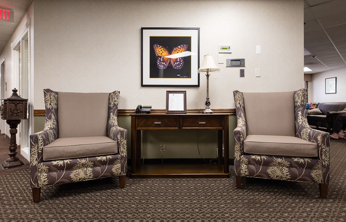 A cozy waiting room featuring two upholstered chairs, a table with a lamp, and butterfly artwork on the wall.