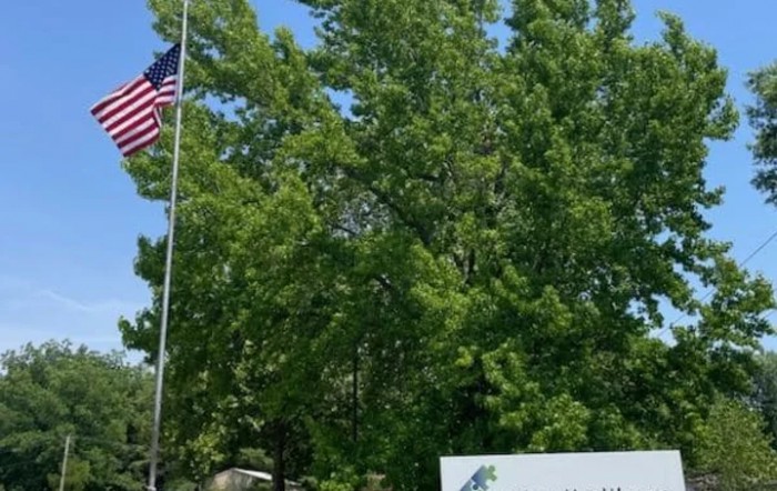 An American flag flies next to a sign surrounded by lush green trees under a clear blue sky.