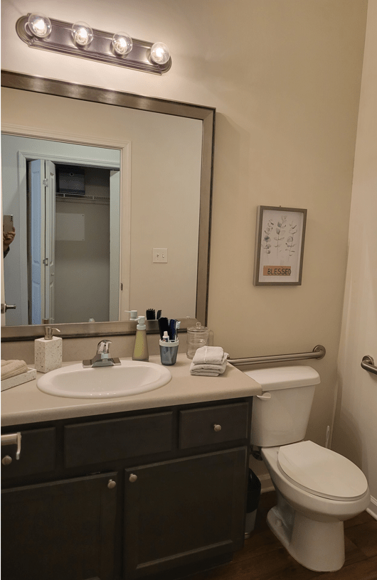 A tidy bathroom with a large mirror above the sink, neatly arranged toiletries, and a small piece of wall art.