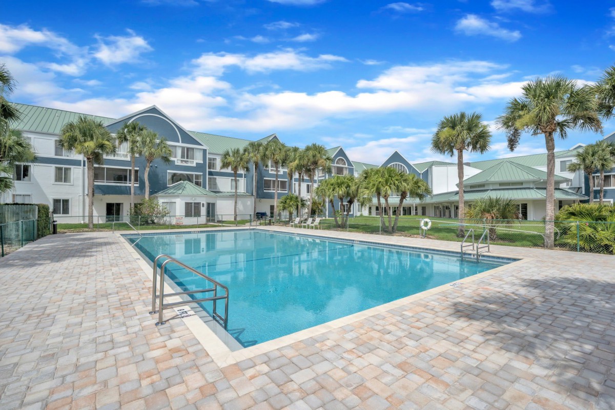 A serene residential area featuring a large outdoor swimming pool surrounded by palm trees and modern buildings under a clear blue sky.