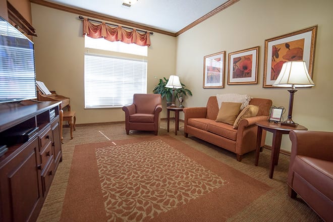 A cozy living room with a sofa, armchair, and a decorative rug, featuring framed artwork and a large window.