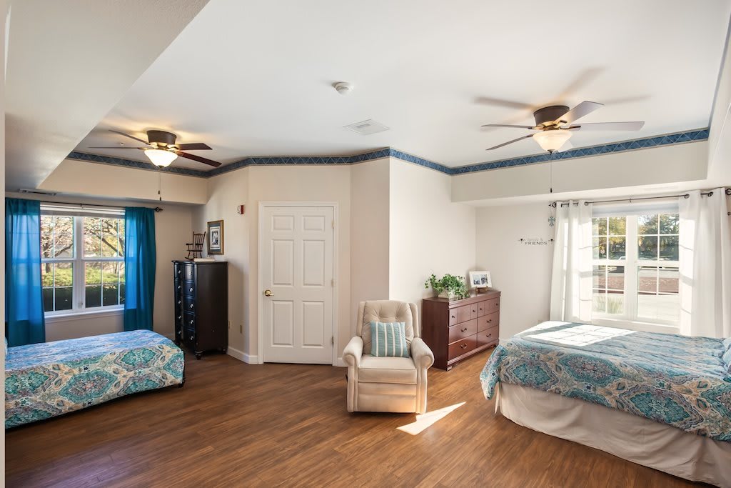 A spacious bedroom with wooden flooring, featuring a bed, armchair, dresser, and ceiling fans. The room is well-lit with natural light from the windows.