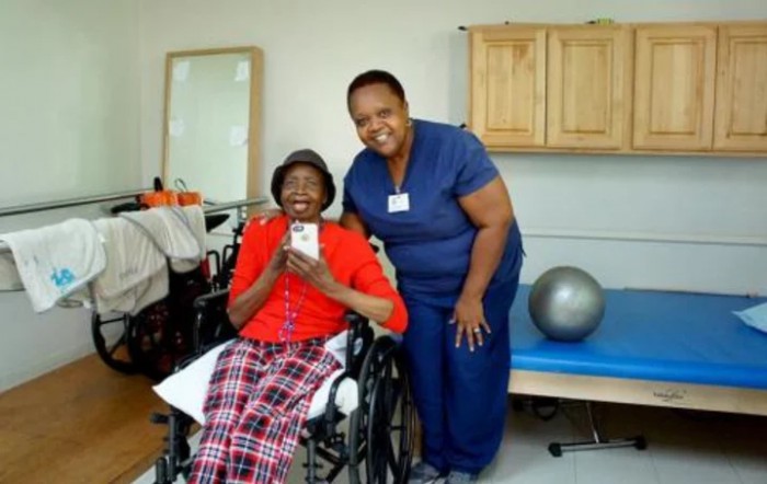 An elderly woman in a wheelchair smiling alongside a caregiver in a healthcare setting.