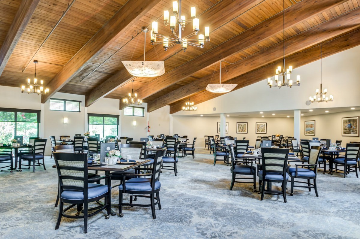 A spacious and elegantly designed dining area with multiple tables and chairs, featuring a wooden ceiling and chandeliers.