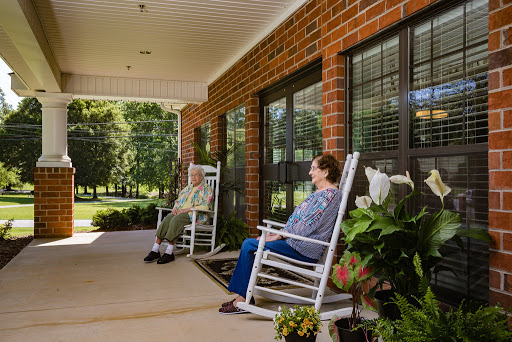 Two individuals enjoy a peaceful afternoon on a porch, seated in rocking chairs surrounded by greenery.