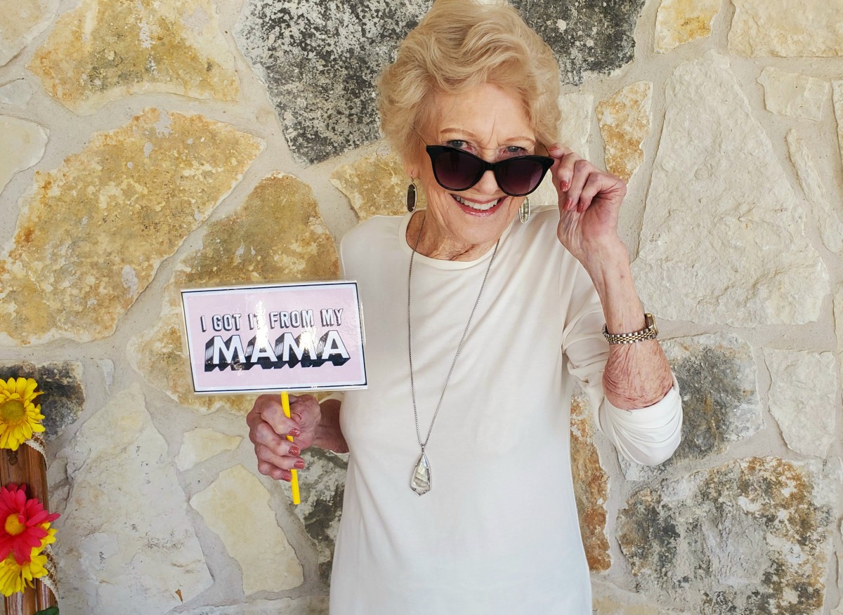 A cheerful woman wearing sunglasses holds a sign while smiling.