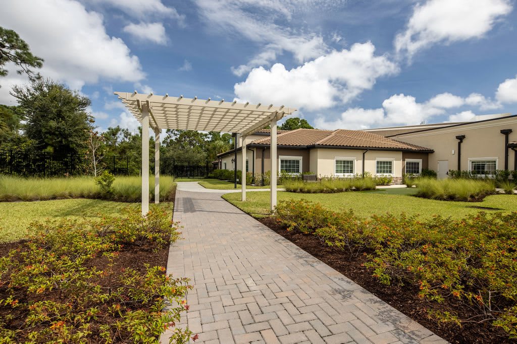 A scenic pathway with a pergola leads to a modern building surrounded by lush greenery under a partly cloudy sky.