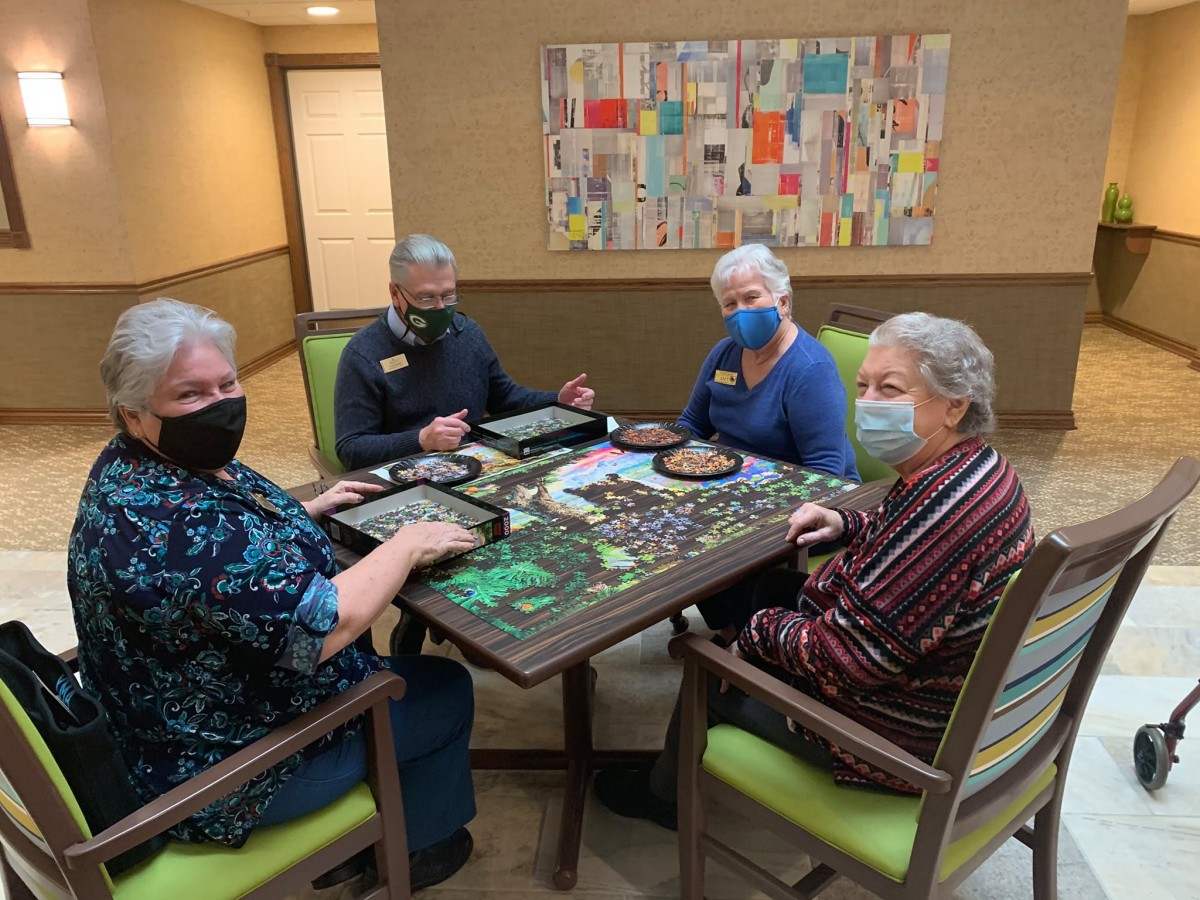A group of seniors wearing masks are gathered around a table, working on a jigsaw puzzle.
