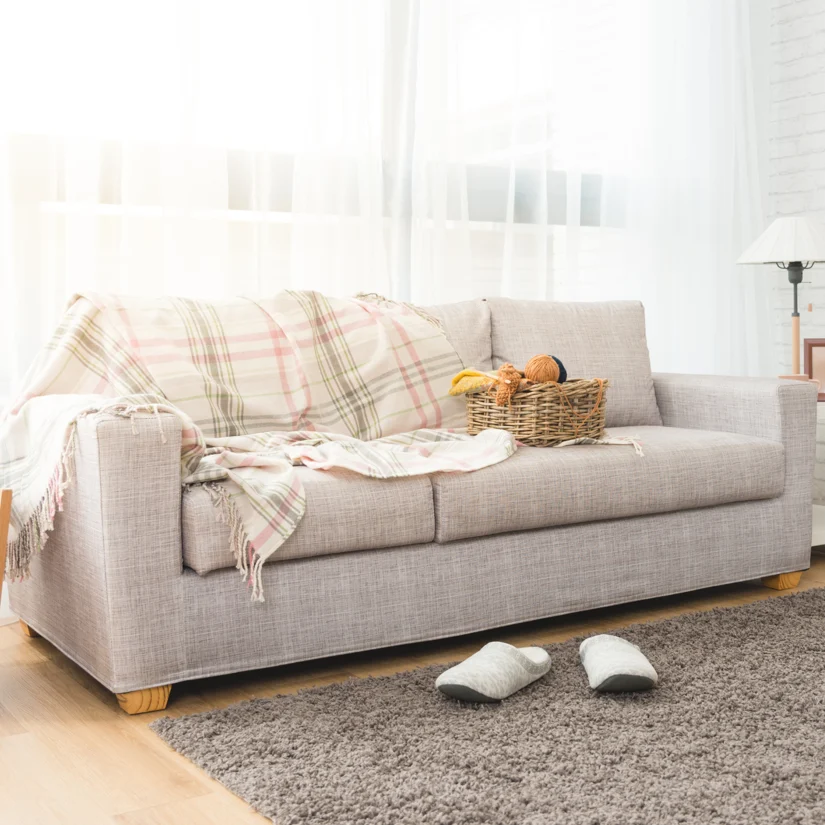 A cozy living room setup featuring a neutral-toned sofa adorned with a plaid blanket and decorative pillows. A pair of slippers rest on the carpet, inviting relaxation.