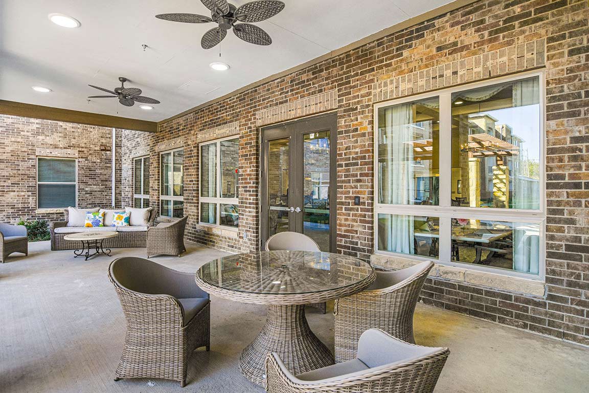 A cozy outdoor patio area featuring wicker furniture and a glass table, set against a brick wall with ceiling fans overhead.