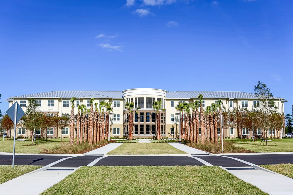 A modern three-story building with palm trees lining the entrance under a clear blue sky.