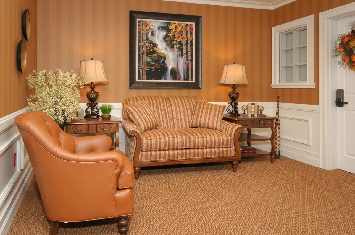 A cozy living room with warm tones, featuring a striped sofa, leather armchair, table lamps, and a landscape painting.