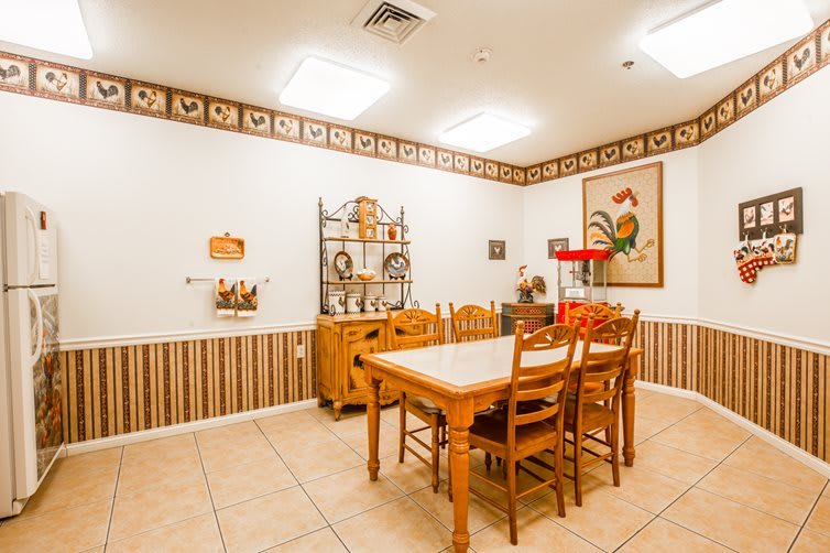 A cozy kitchen dining area with a rustic rooster-themed decor, featuring wooden table and chairs, bright lighting, and decorative artwork.