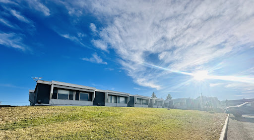 A serene view of a building under a bright blue sky with soft clouds and sunlight illuminating the grassy area.