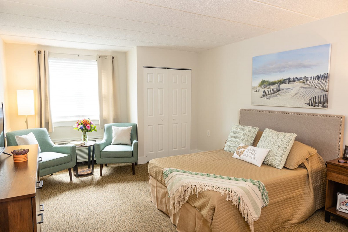 A cozy bedroom featuring a comfortable bed with pillows and a blanket, two armchairs by the window, and a decorative painting on the wall.