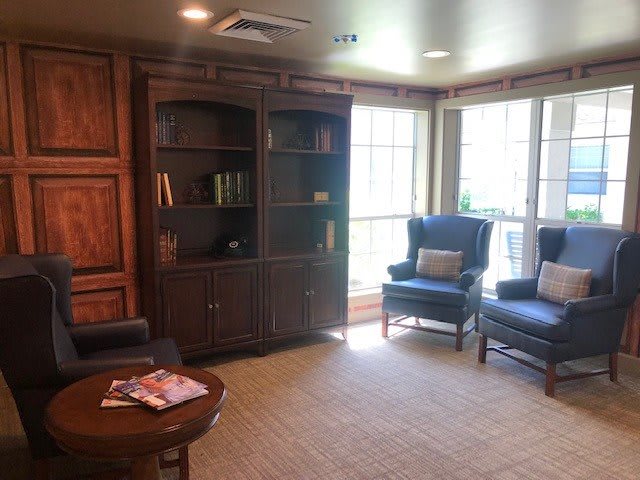 A cozy living room with a bookshelf, two armchairs, and a coffee table near large windows.