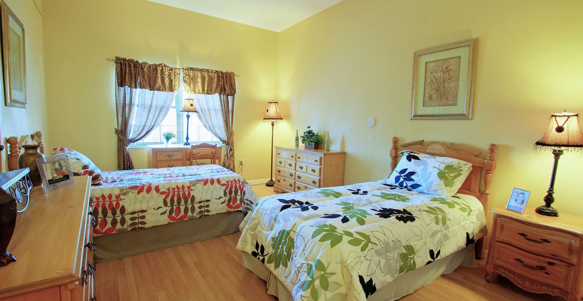A cozy bedroom featuring twin beds with floral bedding, warmly lit by lamps and softened by drapes over the window.