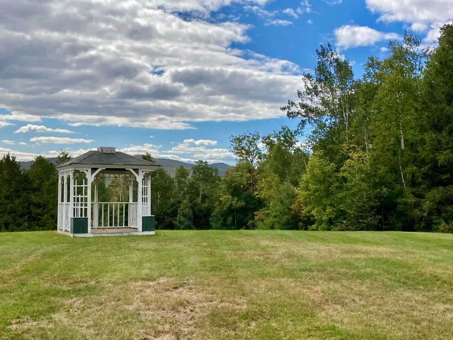 A serene landscape featuring a white gazebo surrounded by lush greenery under a partly cloudy sky.