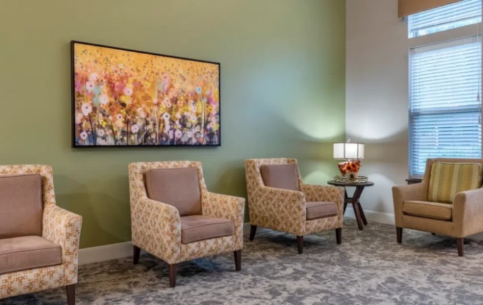A cozy living room setup featuring two patterned armchairs, a side table with a lamp and flowers, and a vibrant painting on the wall.