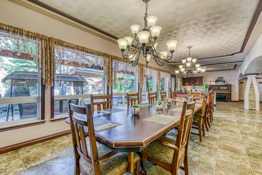 A spacious dining room featuring a large wooden table, multiple chairs, and elegant chandeliers, with large windows offering natural light.