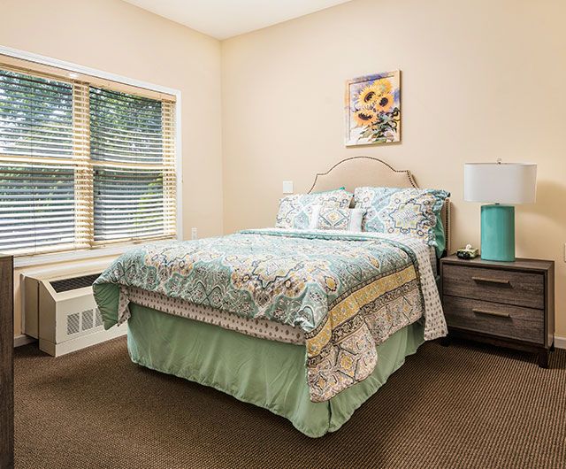 A cozy bedroom featuring a neatly made bed with patterned bedding, a window with blinds, and a nightstand in the corner.