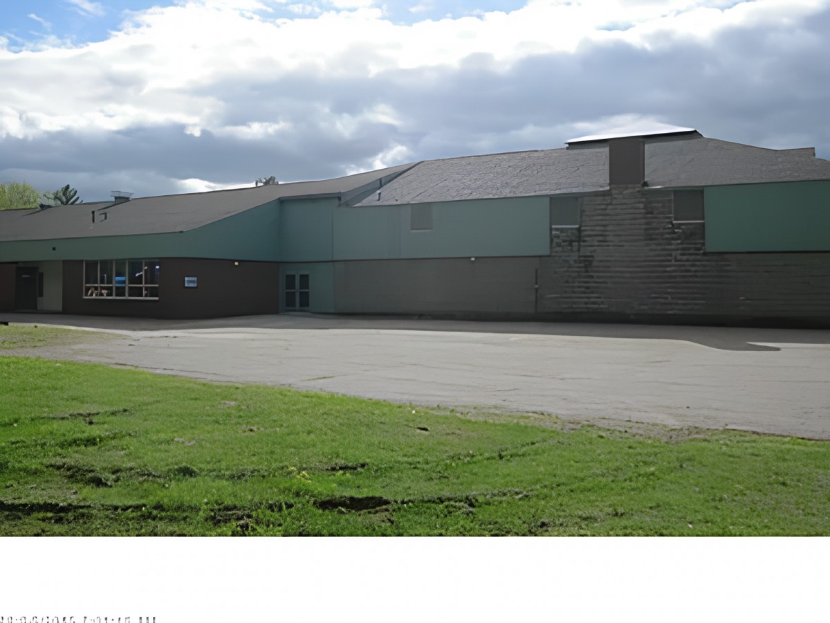 A view of a school building with a paved playground and grassy area under a cloudy sky.
