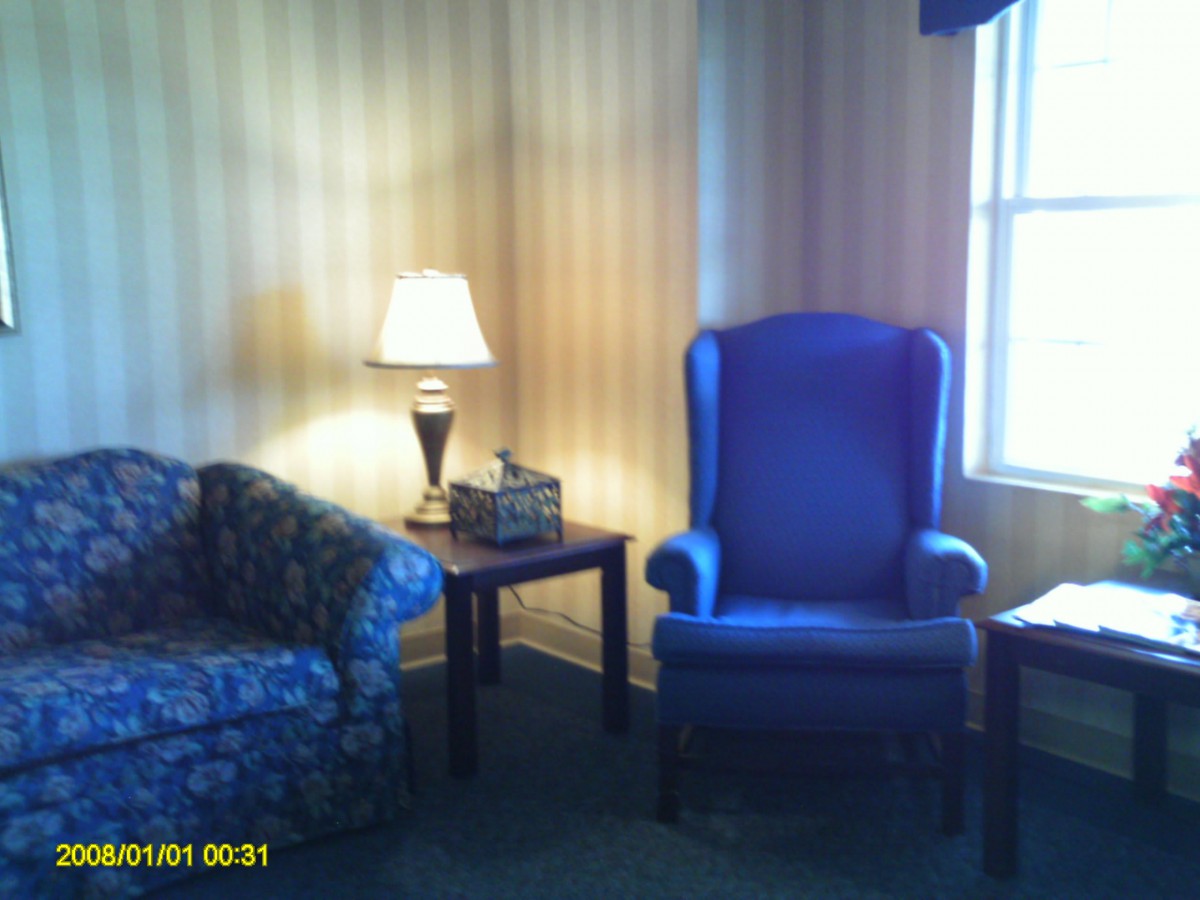 A cozy corner of a living room featuring a blue chair and a patterned couch beside a lamp on a small table.