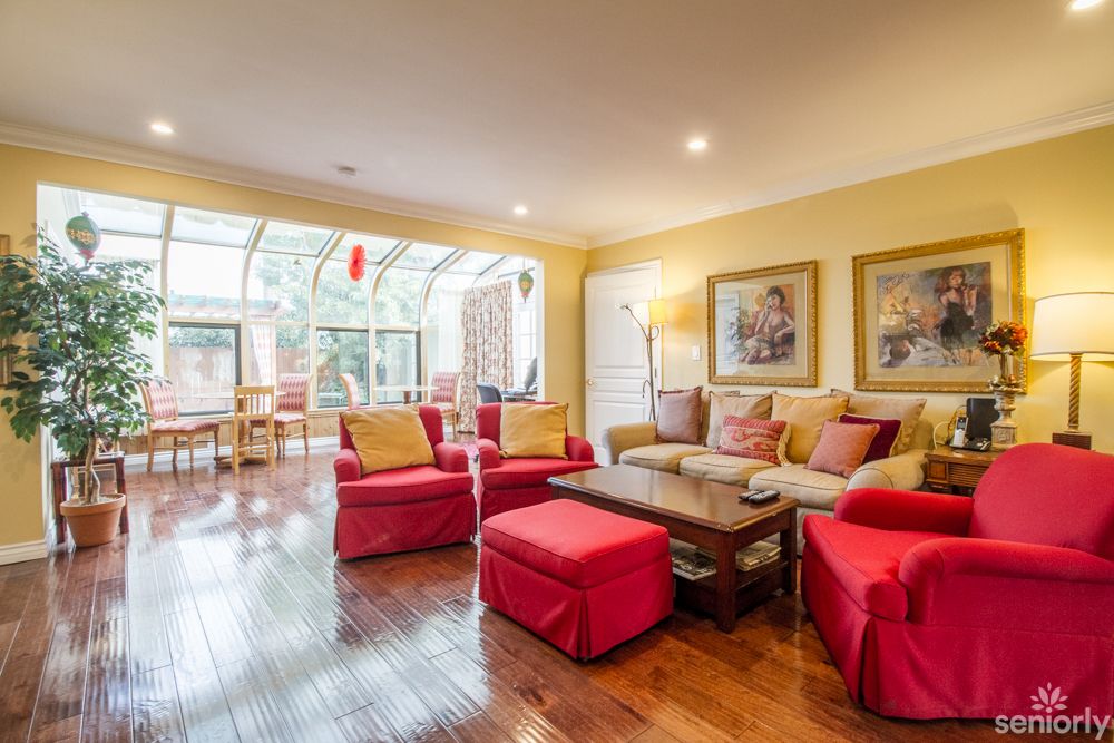 A cozy living room with red chairs, wooden flooring, and large windows leading to a sunroom.