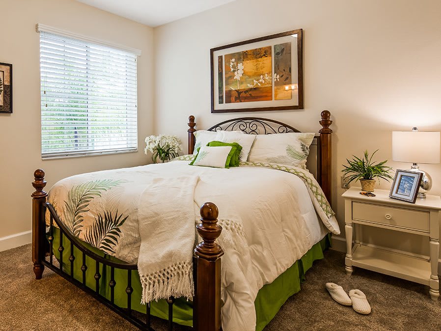 A cozy bedroom with a well-made bed, artwork on the wall, and a nightstand adorned with a plant and lamp.