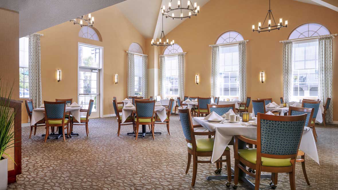 A spacious and elegant restaurant dining area with tables set for service, illuminated by large windows and chandeliers.