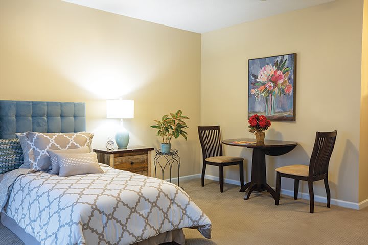 A cozy bedroom with a bed, bedside table, lamp, small round dining table with two chairs, decorative plants, and a floral artwork on the wall.