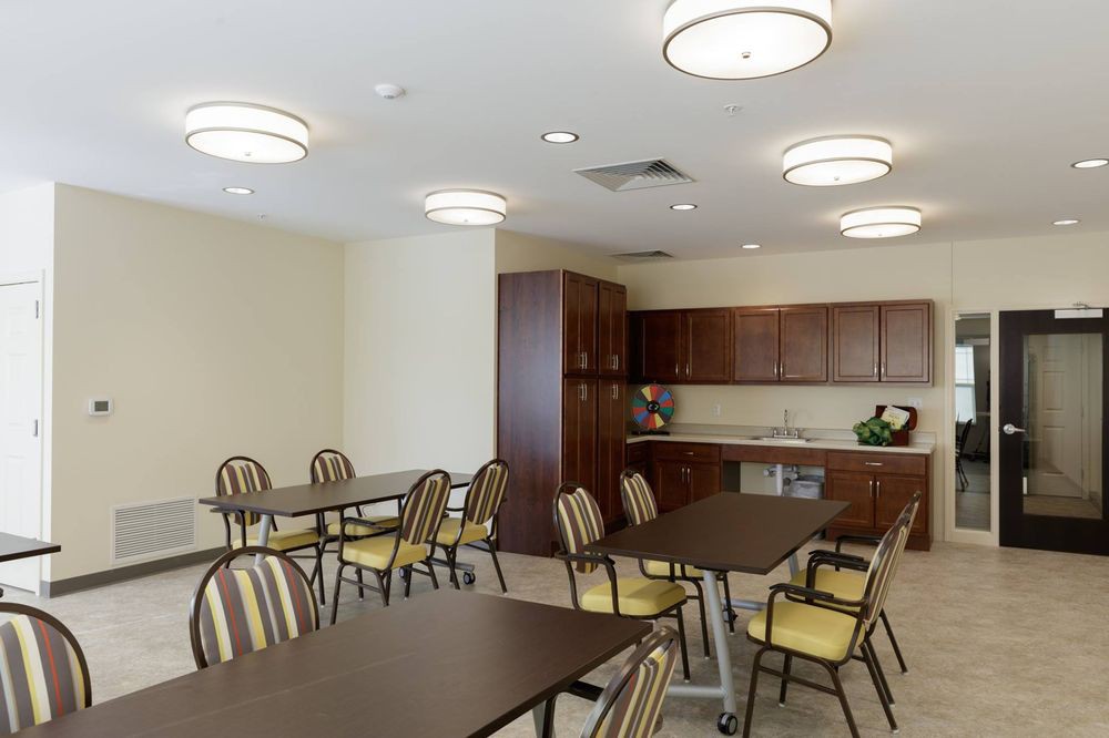 A modern dining room with tables and chairs, featuring overhead lighting and a kitchenette in the background.