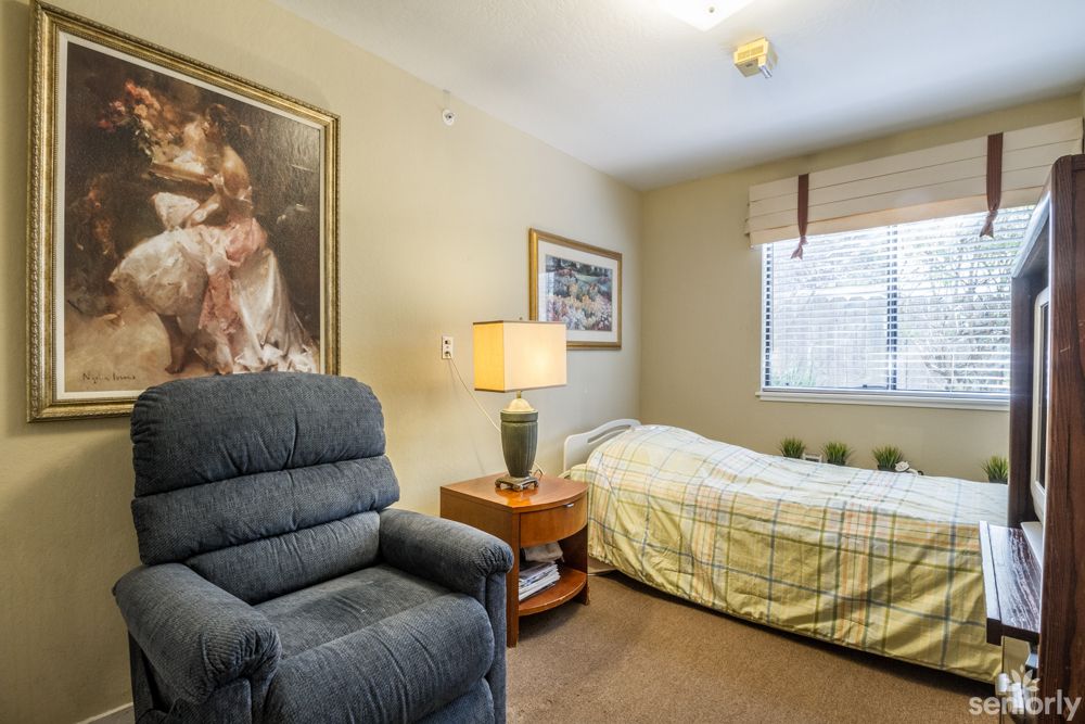 A cozy bedroom with a bed, armchair, and side table featuring a lamp and paintings on the walls.