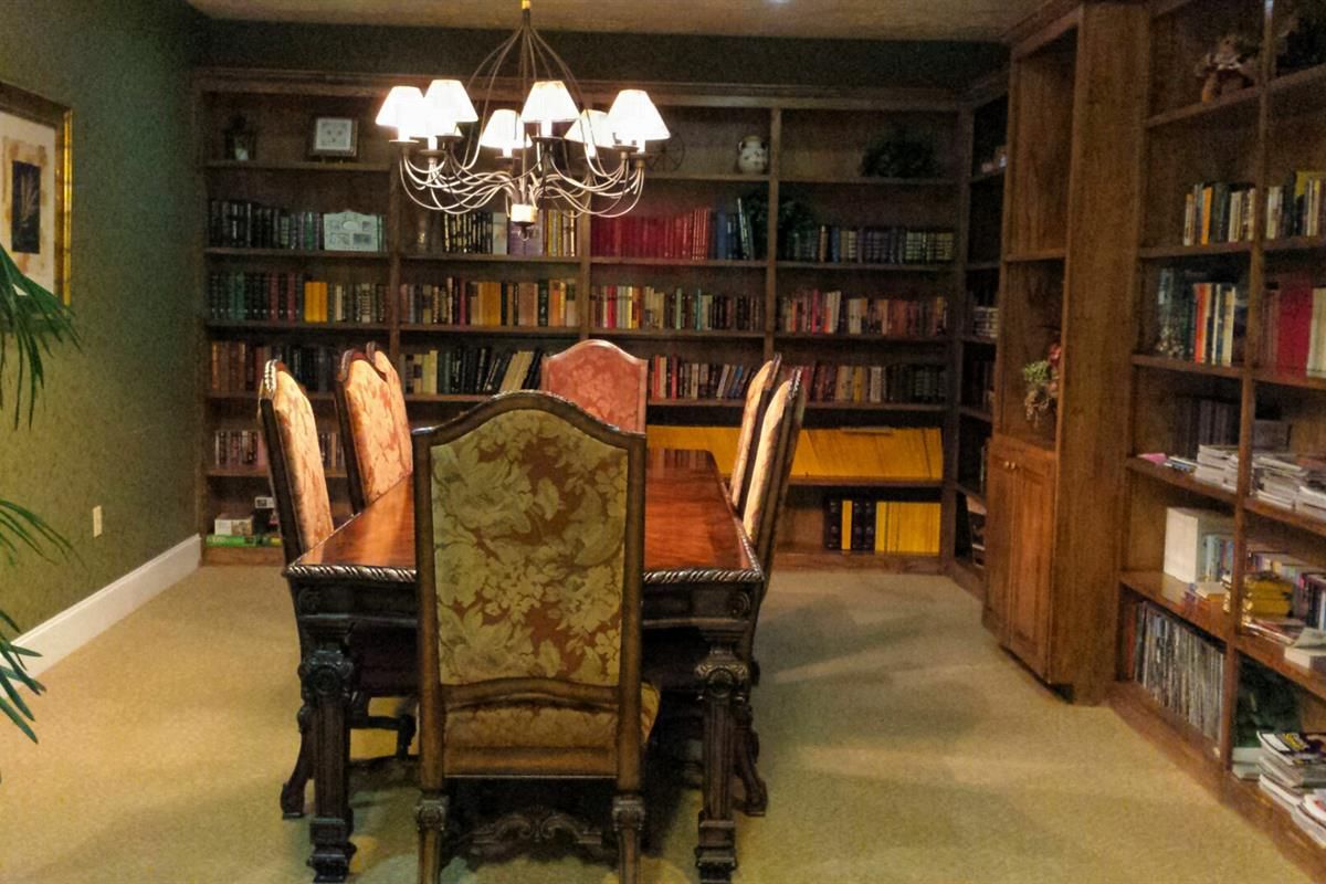 A cozy library room with bookshelves, a wooden table, and elegant chairs under a chandelier.
