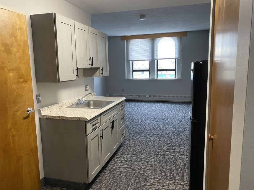 A small kitchen area with cabinets, a sink, and a countertop, featuring two windows in the background.