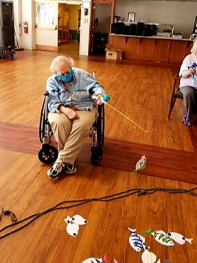 Two elderly individuals engaging in an activity at a care facility, with one using a wheelchair and wearing a mask.