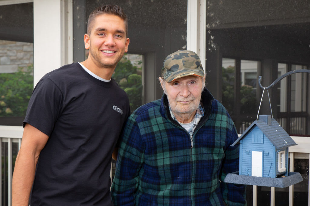 A young adult standing next to an elderly man wearing a plaid jacket and camo hat.