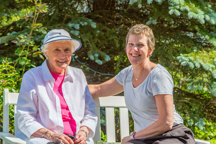 Two women sitting on a bench, smiling at the camera in an outdoor setting.