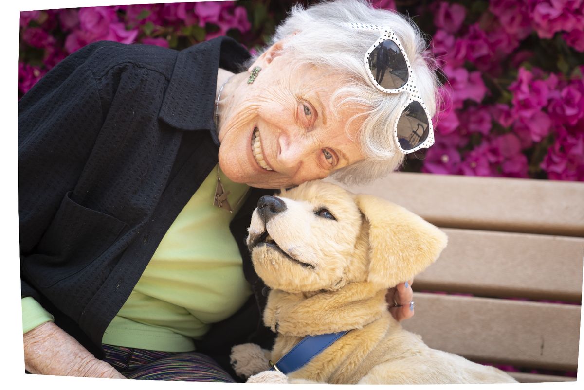 An elderly woman smiling at a dog while sitting on a bench outdoors.